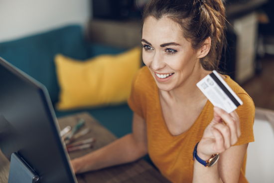 Junge Frau an einem Laptop, hält mit einer Hand eine Bankkarte in die Luft